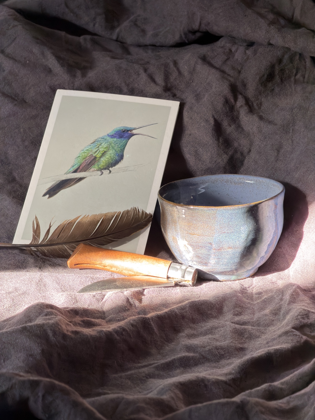 Keramiktasse, Feder und Vogelillustration auf dunklem Leinen im Herbstlicht